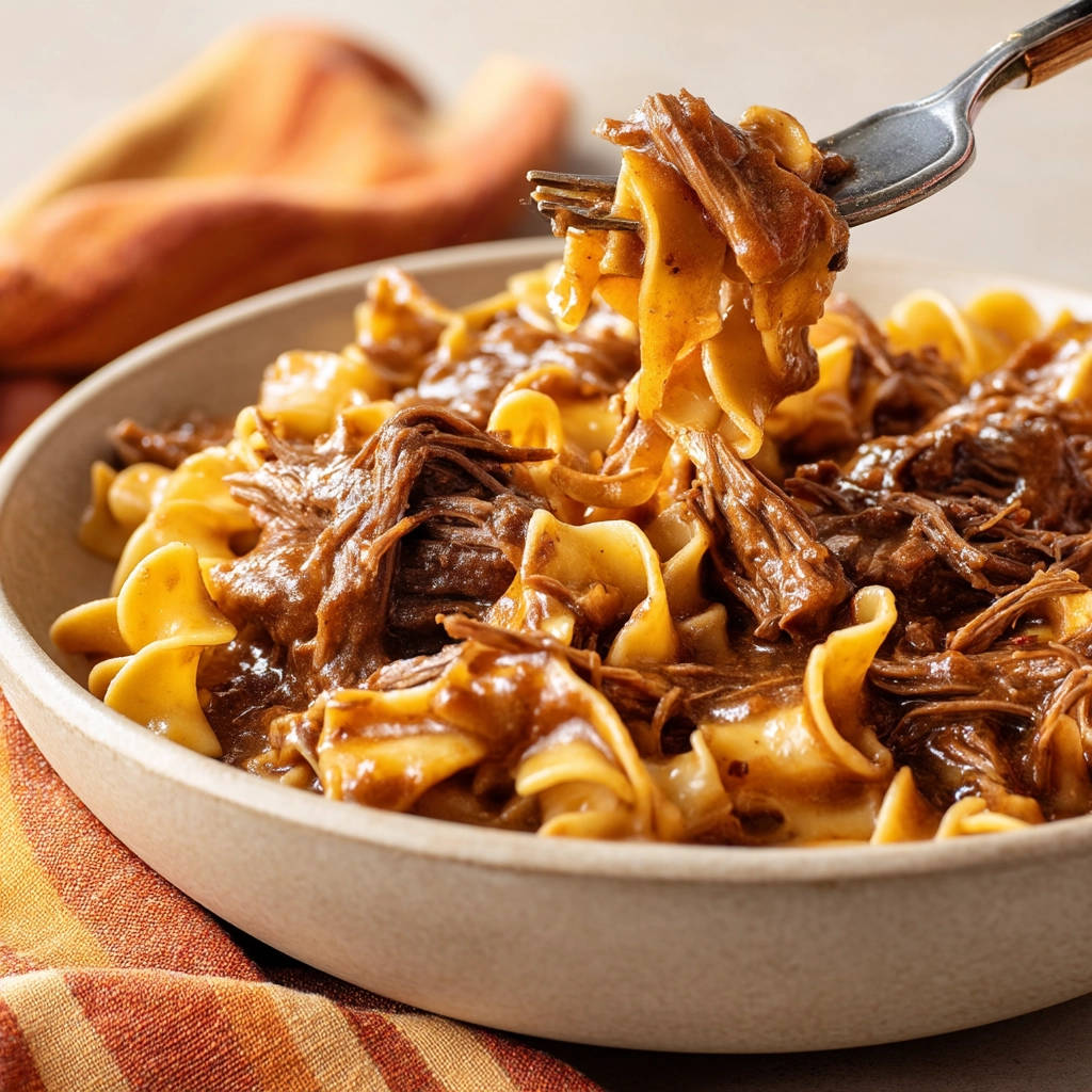Comforting beef and noodles with a forkful being lifted, showcasing tender shredded beef and wide egg noodles in rich gravy.